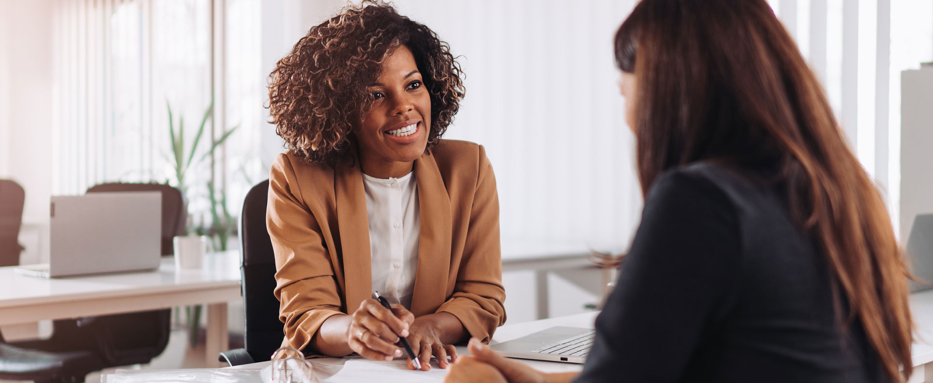 Two women talking together at a desk in an office setting.